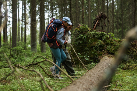 Middle aged Caucasian man and young adult Black woman hiking together in forest, carrying backpacks...