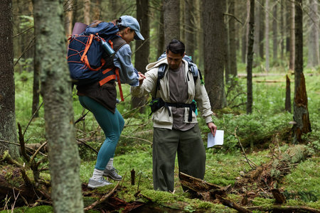Young adult Caucasian man helping young adult Caucasian woman cross fallen tree while hiking in...