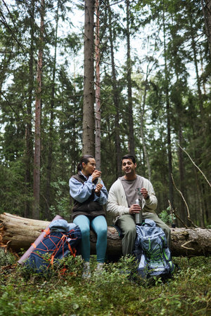 Young adult woman and young adult man sitting on fallen tree in forest, eating snack and drinking from thermos during hiking trip with backpacks on ground nearbyの写真素材