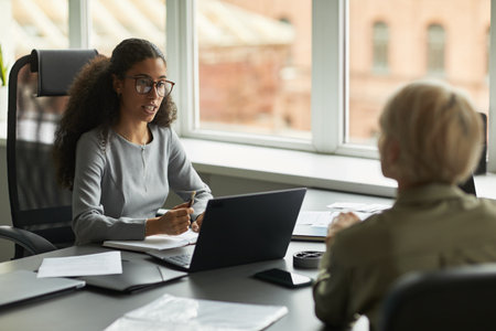 Young adult woman sitting at desk talking to middle aged woman during business meeting, holding pen and making notes, laptop and documents on tableの写真素材