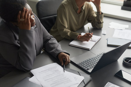 Black woman and Caucasian woman sitting at desk working with documents and laptop, Black woman holding head appearing stressed while Caucasian woman writing notes in notebookの写真素材