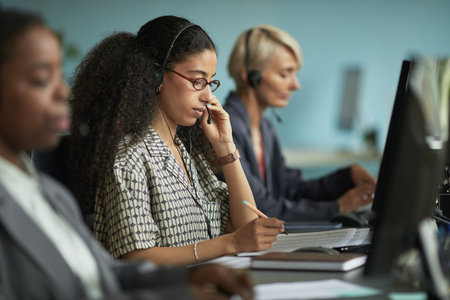 Young adult woman wearing headset working at computer in modern office, sitting at desk with colleagues including middle aged woman using headset in backgroundの写真素材