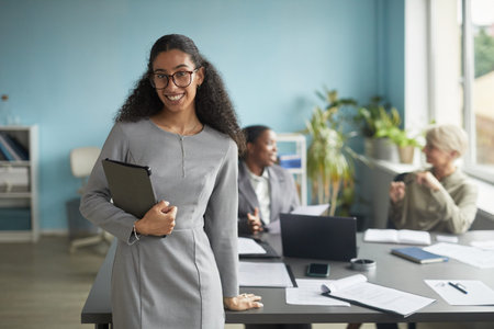 Portrait of young adult biracial woman smiling at camera while standing in modern office holding...