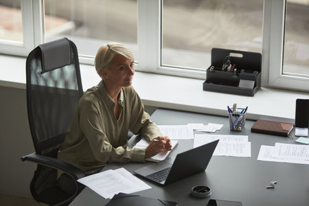Caucasian middle aged woman sitting at desk working on laptop surrounded by documents in modern office, hands clasped, looking thoughtfully out window during business dayの写真素材