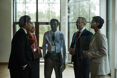 Group of middle aged multiethnic business professionals standing in office discussing project, including Caucasian man, woman, Black man, wearing corporate badgesの写真素材