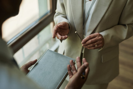 Middle aged Caucasian woman holding pen and gesturing while standing near window, interacting with middle aged man holding notebook, both engaged in business discussion indoorsの写真素材