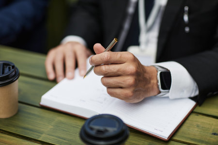 Unrecognizable businessman writing in notebook with pen, wearing suit and smartwatch, sitting at...