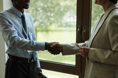 Caucasian middle aged woman and Black man shaking hands in office setting, both wearing business attire, engaging in professional agreement near large windowの写真素材