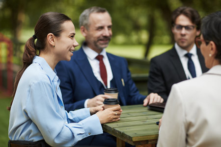Caucasian young woman and middle aged men sitting outdoors at wooden table holding coffee cups, engaging in business conversation, talking and smiling during lunch breakの写真素材
