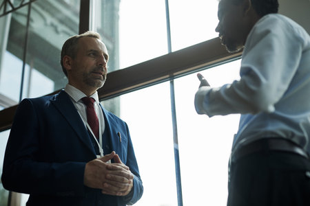 Caucasian middle aged man standing with hands clasped listening to Black young businessman gesturing...