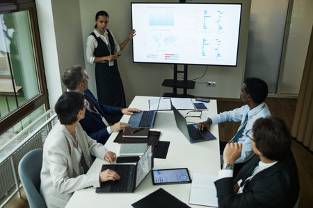 Young Caucasian woman presenting data charts on large screen to group of middle aged multiethnic men...