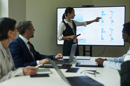 Young woman presenting data charts on digital screen to group of middle aged multiethnic business...