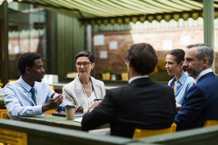 Group of middle aged and young adult multiethnic professionals sitting outdoors at cafe discussing...