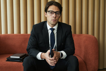 Portrait of pensive young Caucasian man wearing glasses sitting on sofa holding smartphone with both...
