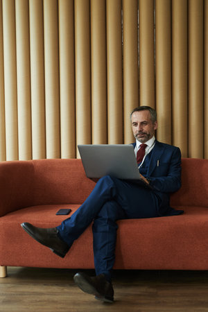 Middle aged Caucasian man sitting on sofa using laptop, crossing legs and focusing on screen,...