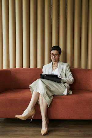 Busy middle aged Caucasian woman in suit sitting on sofa working on digital tablet, wearing glasses, focusing on screen, legs crossed, vertical wall decor in backgroundの写真素材