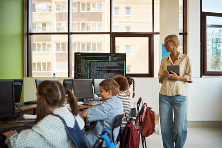 Caucasian middle aged woman holding tablet standing near group of diverse children sitting at desks...