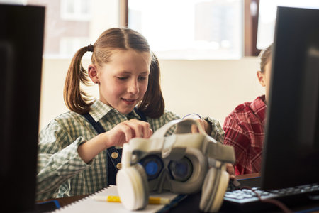 Caucasian girl child assembling virtual reality headset at desk in classroom, sitting between...