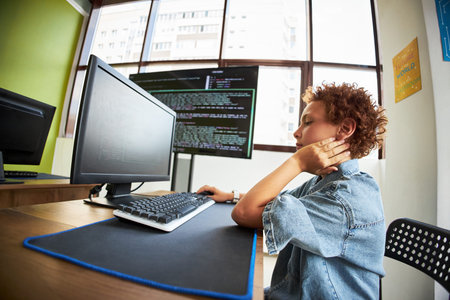 Teenager boy sitting at desk using desktop computer, holding neck with one hand, looking at monitor...