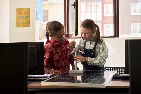 Two Caucasian girls working together assembling wind turbine model on solar panel in classroom, focusing on renewable energy project, both appearing as children around elementary school ageの写真素材
