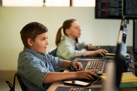 Caucasian boy sitting at desk using computer mouse and keyboard in classroom, focusing on monitor,...