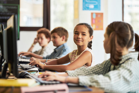 Group of preteen Caucasian and multiethnic children sitting at desks using desktop computers in classroom, girl in foreground turning and smiling at classmate while typing on keyboardの写真素材