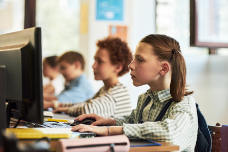 Caucasian girl child sitting at desk using desktop computer in classroom, focusing on screen while other children working in background, participating in educational activityの写真素材