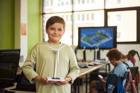Caucasian boy holding wind turbine model standing in classroom, smiling at camera while classmates working on computers in background, STEM education project in progressの写真素材