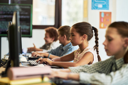 Group of Caucasian and Black children sitting at desks using desktop computers in classroom, focusing on typing and learning coding skills, teacher visible in background assisting studentsの写真素材