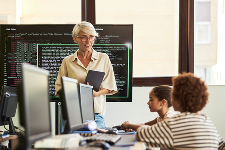 Middle aged Caucasian woman teaching coding class, holding digital tablet while standing near large...