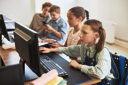 Group of Caucasian and multiethnic children sitting at desk using desktop computers and smartphone...