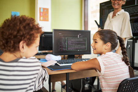 Caucasian girl and Black boy sitting at desk using computer in classroom, smiling and talking to...