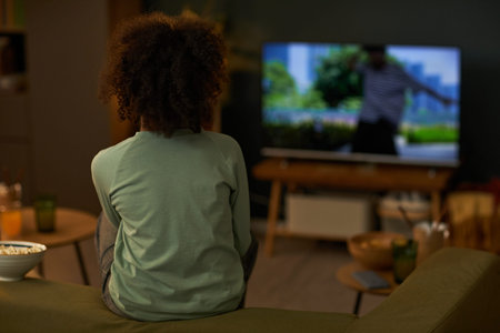 Black child sitting on sofa watching television screen showing outdoor scene, back facing camera,...