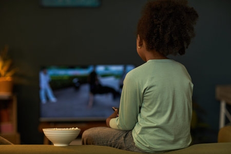 Black child sitting on sofa watching television with bowl of popcorn on table in foreground, holding...