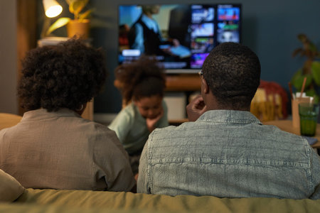 Black woman and Black man sitting on couch watching television while Black child playing on floor in...