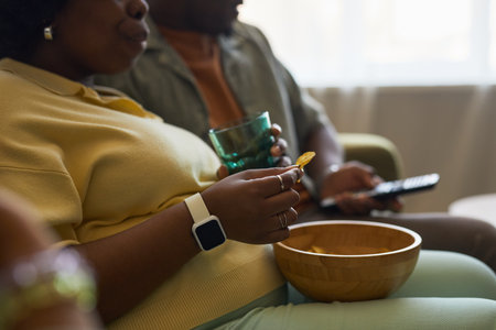 Woman eating snacks from wooden bowl while sitting beside man holding glass and remote control, both relaxing together on sofa in casual home settingの写真素材