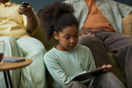 Girl child sitting between adults using digital tablet, focusing on screen while adult hands holding...