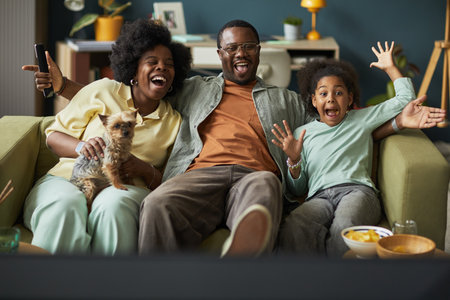 Black woman, Black man, and Black child sitting on sofa watching television together, smiling and cheering with excitement, woman holding small dog, snacks visible on table in foregroundの写真素材