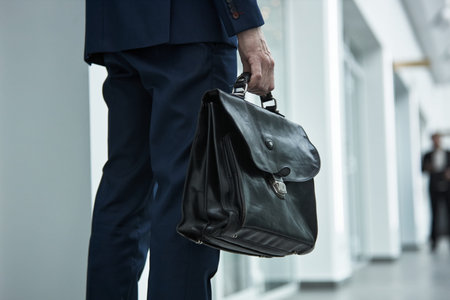 Anonymous businessman in suit holding leather briefcase walking through modern office corridor,...
