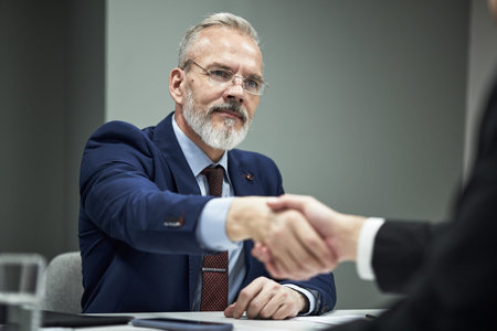 Caucasian middle aged male executive with gray beard shaking hands with partner business meeting,...