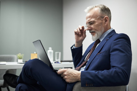 Senior Caucasian man sitting at desk using laptop, adjusting eyeglasses while focusing on screen, business professional working in modern office environmentの写真素材