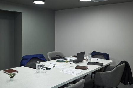 Modern office meeting room showing empty chairs around conference table, suit jackets, open laptop,...