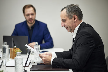 Mature Caucasian man reading documents during business meeting with colleague working on laptop in...
