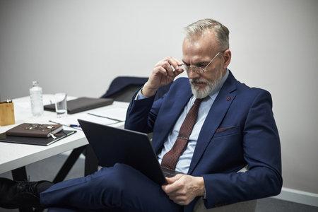 Middle aged Caucasian man with gray beard sitting at desk using laptop, adjusting eyeglasses while concentrating on screen, business documents and water bottle visible on tableの写真素材