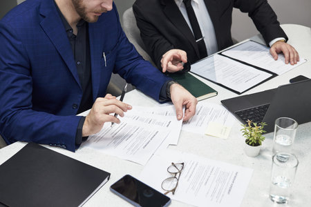 Two male managers in formal suits discussing documents at desk, reviewing printed contracts and...