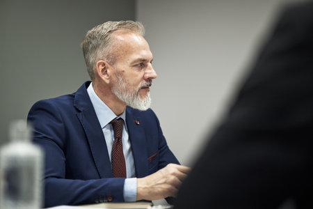 Senior Caucasian man sitting at table participating in business meeting, wearing formal suit, looking attentively to side, engaging in professional discussion with colleaguesの写真素材