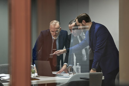 Three middle aged Caucasian male managers reviewing business contract on laptop in modern office, one man pointing at document, business teamwork and discussion, behind glass wallの写真素材