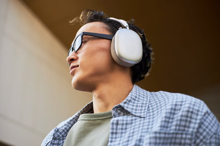 Young adult Asian man wearing over ear headphones and glasses looking away from camera outdoors,...