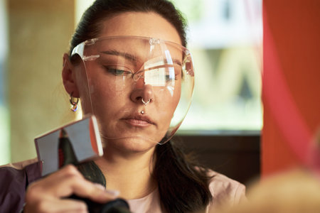Young adult Caucasian woman wearing protective face shield using handheld device, focusing intently on task, close up of face and upper body, working in professional environmentの写真素材