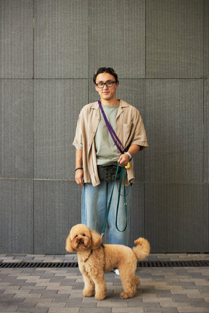 Portrait of young adult Asian man standing and holding dog leash, looking at camera with curly...